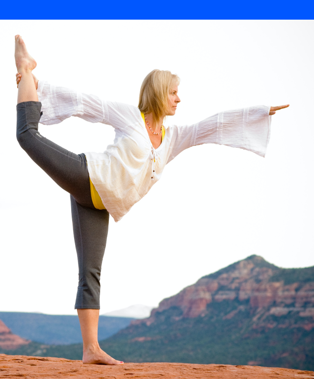 A lady performing a yoga pose on a rocky ground with a scenic mountain backdrop, wearing a white flowing top, grey leggings, and barefoot, with one leg lifted and arms extended to the sides.