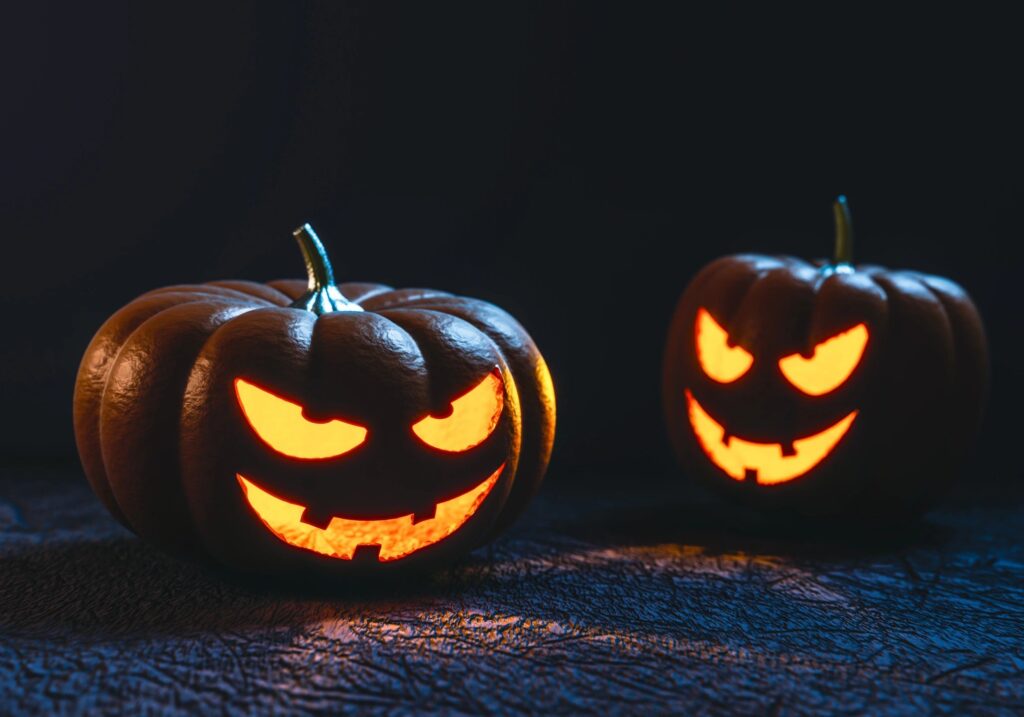 Two carved pumpkins with glowing eyes and sinister smiles illuminated from within, sitting on a dark textured surface with a black background.
