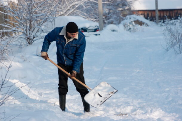 A person in winter clothing shoveling snow with a large shovel on a snow-covered path surrounded by snowy bushes and trees, with snow-covered vehicles and a wooden structure in the background.