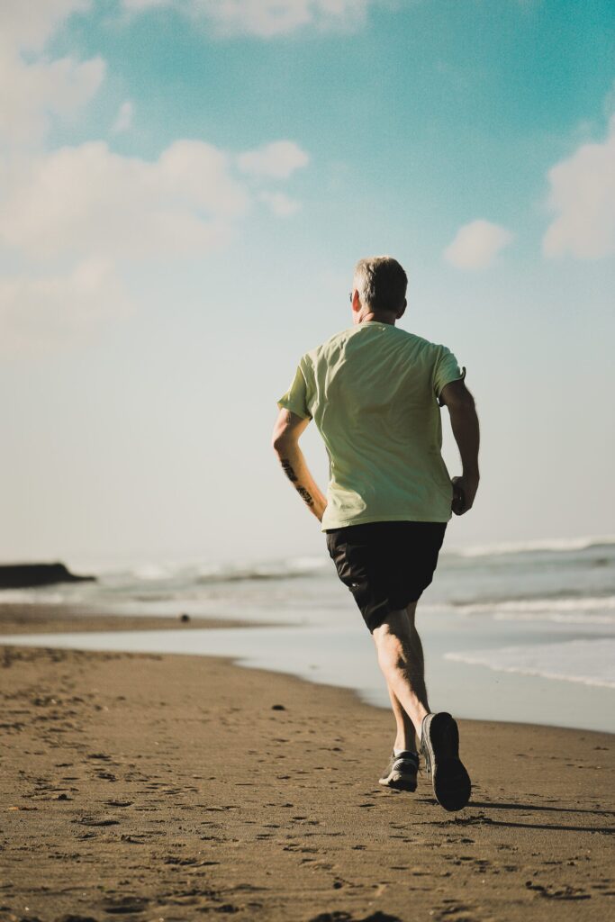A man wearing a light green t-shirt and black shorts running on a sandy beach with the ocean on the right and a clear sky in the background.