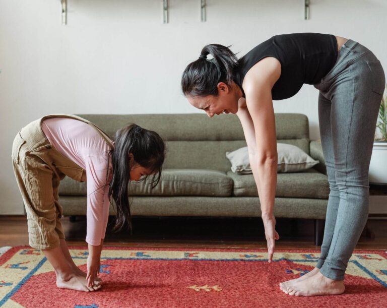 A woman and a young child are bending forward reaching for their toes in a room with a couch and a colorful rug on the floor. The child is wearing a pink top and khaki pants, and the woman is dressed in a black top and gray pants. They both have their hair tied up.