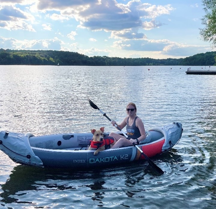 A person in a life vest and a dog wearing a red safety jacket are sitting in an inflatable kayak labeled "INTEX DAKOTA K2" on a body of water. There are trees in the background under a cloudy sky.