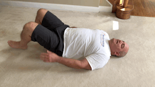 A man lying on his back with legs bent, oscillating knees side-to-side for a pelvic exercise.