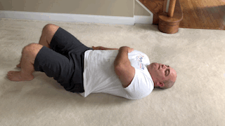 Bald man in white shirt and black shorts lying on his back on a carpeted floor, doing bicycle crunches by lifting his legs and touching elbow to opposite kneealternately.