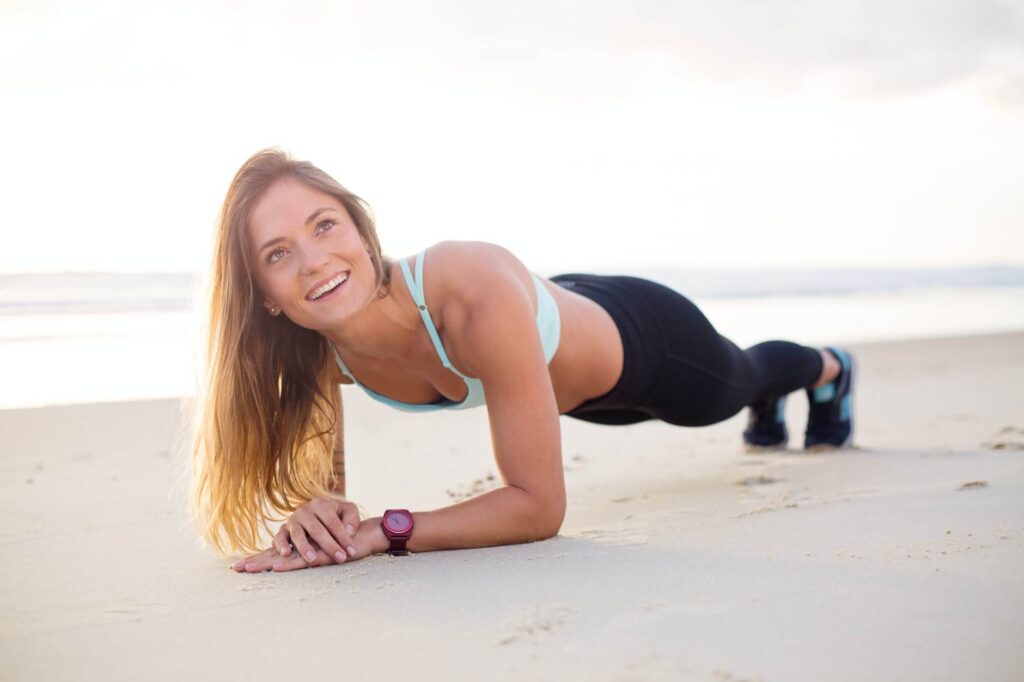 A person wearing a light blue sports bra and black leggings is performing a forearm plank on a sandy beach. They have long blonde hair reaching down the back, and a sport watch on their left wrist. The background shows a calm sea and a clear sky during what appears to be sunrise or sunset.
