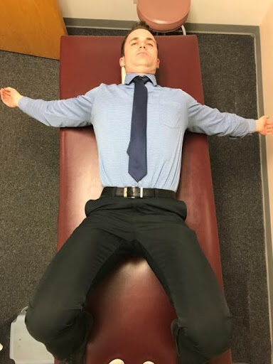 A person lying on their back on a brown medical examination table, wearing a light blue shirt, dark blue tie, black pants, and a belt. Their arms are outstretched, and their legs are slightly apart.