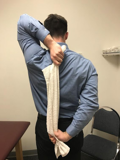 A man in a blue dress shirt and black pants is standing with his back to the camera, reaching over his shoulder with one hand to hold a white towel draped down his back.