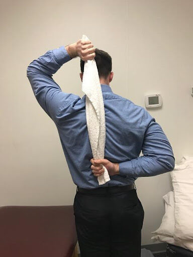 A man in a blue dress shirt and black pants is standing with his back to the camera, reaching over his shoulder with one hand to hold a white towel draped down his back.