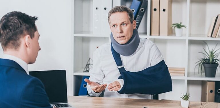 Two men in business attire sitting at a desk with a laptop between them, with one man wearing a neck brace and arm sling gesturing with his hand during a discussion in a modern office environment.