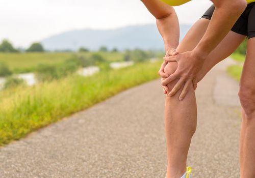 Runner holding their knee in pain on a path with nature in the background.