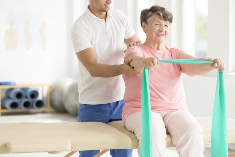 A physical therapist assisting a patient with an exercise using a green resistance band in a bright, well-lit therapy room. The patient is seated while the therapist, dressed in a white polo shirt and blue pants, provides guidance and support from behind.