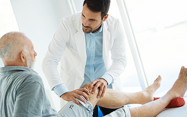 A healthcare professional in a white coat and blue shirt examines the knee of a patient sitting on an examination table in a bright, modern medical office. The patient appears to be an older adult, wearing a gray shirt and shorts, with a supportive red object under the knee, suggestive of a physical therapy session.