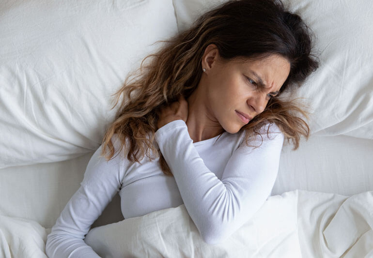 A woman lying on her side on a white bed with white pillows, wearing a white long-sleeved shirt, with her hand resting on her neck.