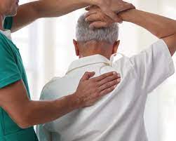 A physical therapist assisting an elderly man with a stretching exercise, holding his arms behind his head and supporting his back.