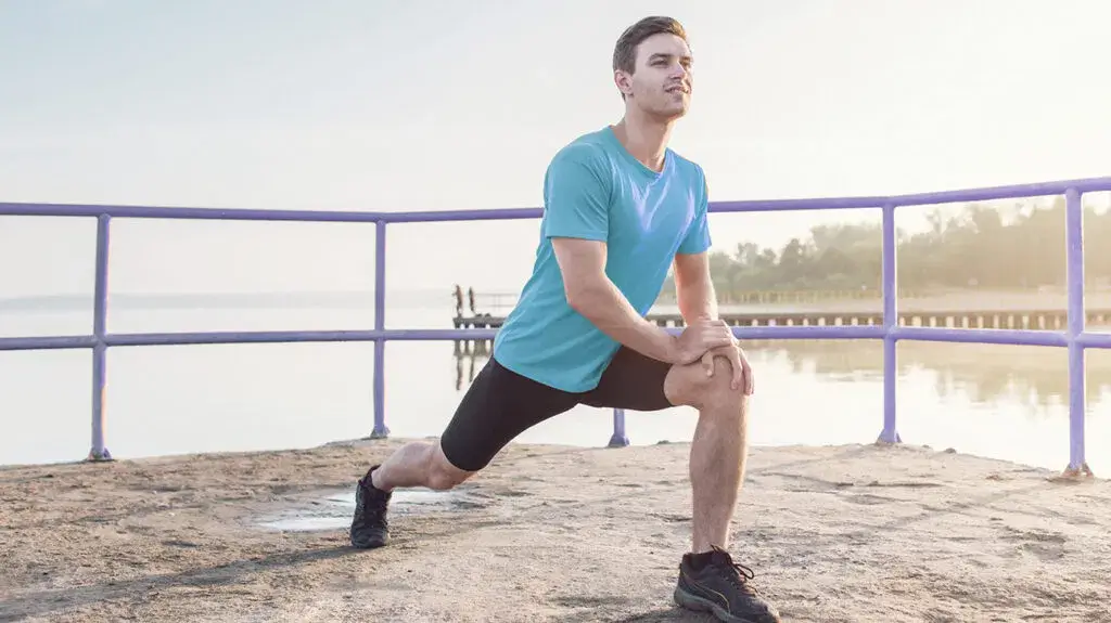 A person in a blue t-shirt and black shorts doing a forward lunge exercise on a concrete surface by a lake, with their hands on their front thigh, a blue railing in the background and a hazy sunrise over the calm water.