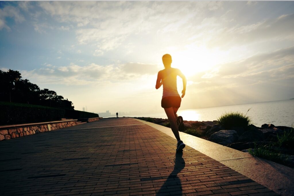 A person running on a paved path along the waterfront with the sun setting in the background.
