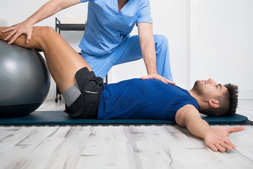 A person in blue scrubs assisting a patient with knee brace lying on a mat performing a physiotherapy exercise using a stability ball.