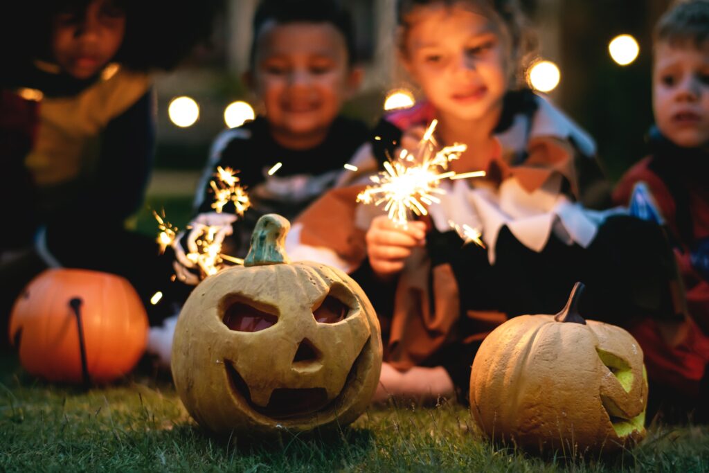 Carved pumpkins on the ground with lit sparklers in the foreground during twilight. The warm glow adds a festive atmosphere likely indicative of Halloween celebrations.
