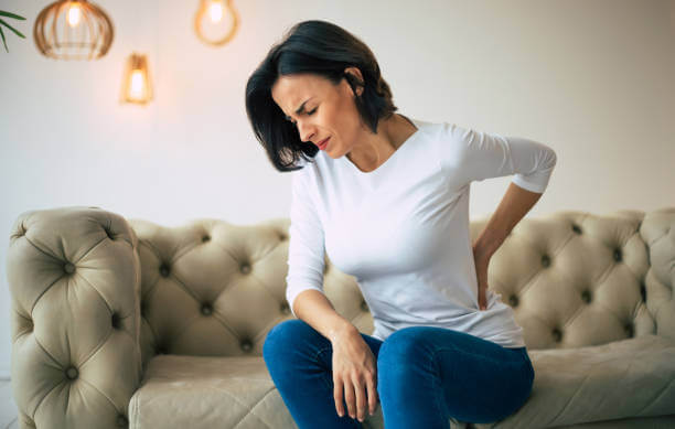 A woman in a white top and blue jeans sitting on a beige tufted sofa holding her lower back as if experiencing pain, with pendant lighting in the background.