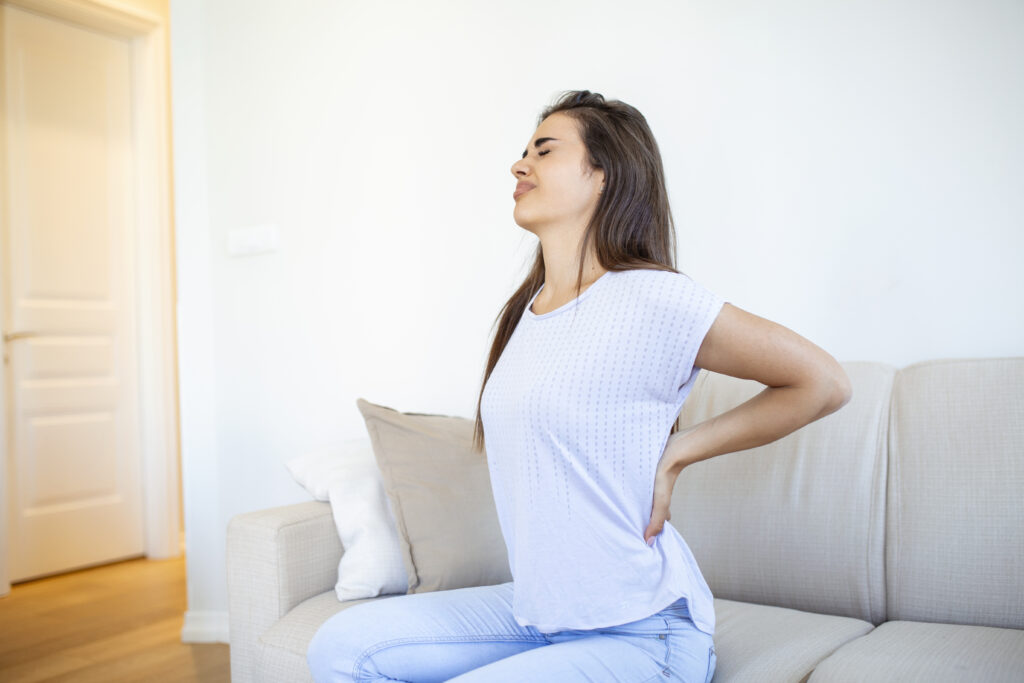 A girl in a white t-shirt and blue jeans sitting on the edge of a beige couch with one hand on their lower back, as if stretching or having back pain, in a bright room with a closed door in the background.
