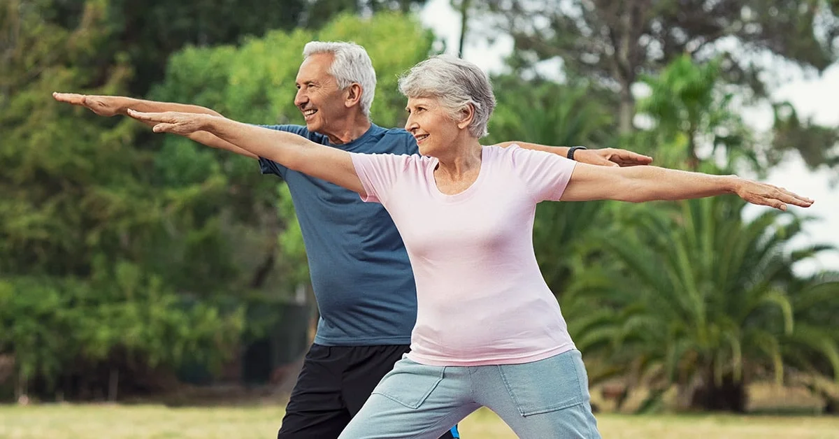Senior couple dressed in sportswear doing stretching exercises in a park with green trees in the background.