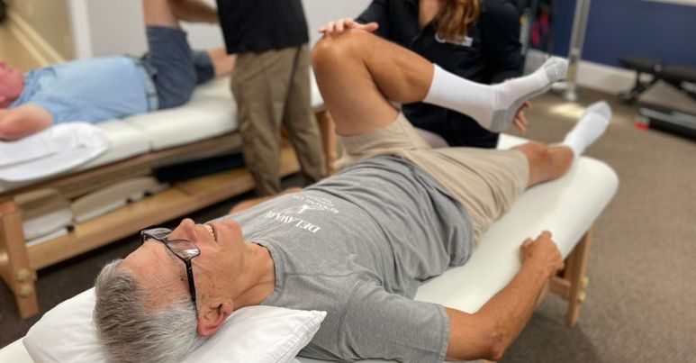 A man lying on a physical therapy table receiving treatment on his leg from a therapist.