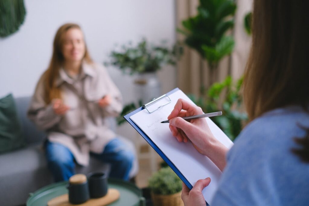 A therapist taking notes on a clipboard while conversing with a female patient seated on a couch in a plant-filled therapy room