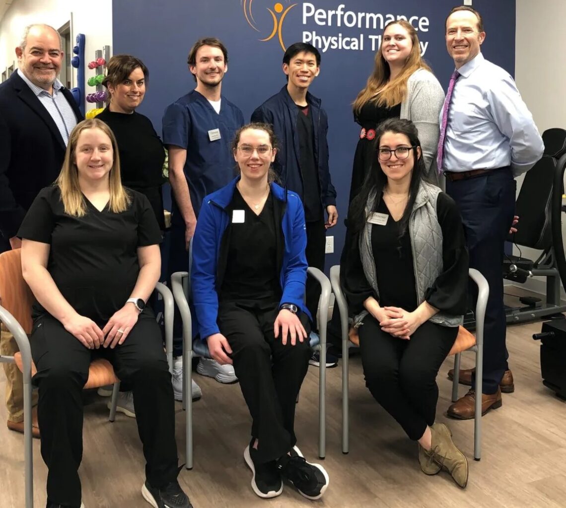 A group of healthcare professionals in dark uniforms pose together in front of a Performance Physical Therapy sign.