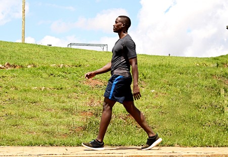 A person walks confidently on a sunny day, wearing athletic clothes, on a wooden path surrounded by green grass and trees.