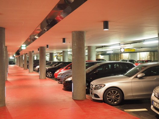 A well-lit underground parking garage featuring multiple parked cars and red flooring, with concrete columns supporting the ceiling.