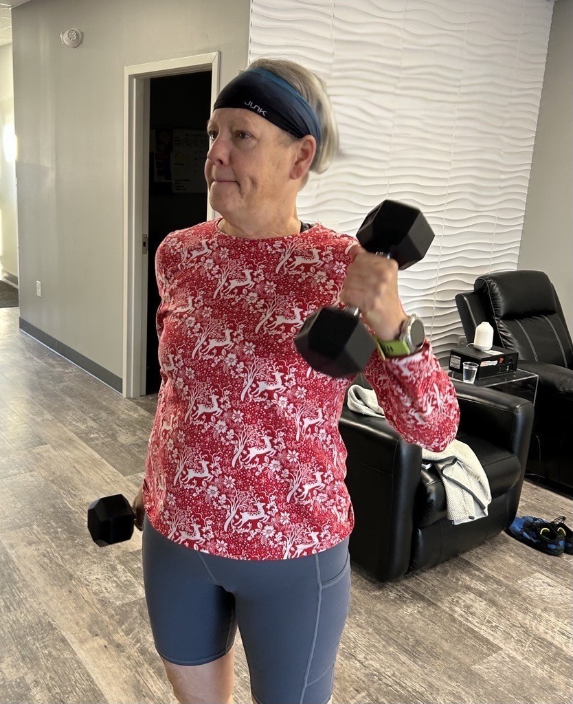 A person wearing a red patterned long-sleeve shirt and gray shorts is lifting dumbbells in a modern, well-lit room.