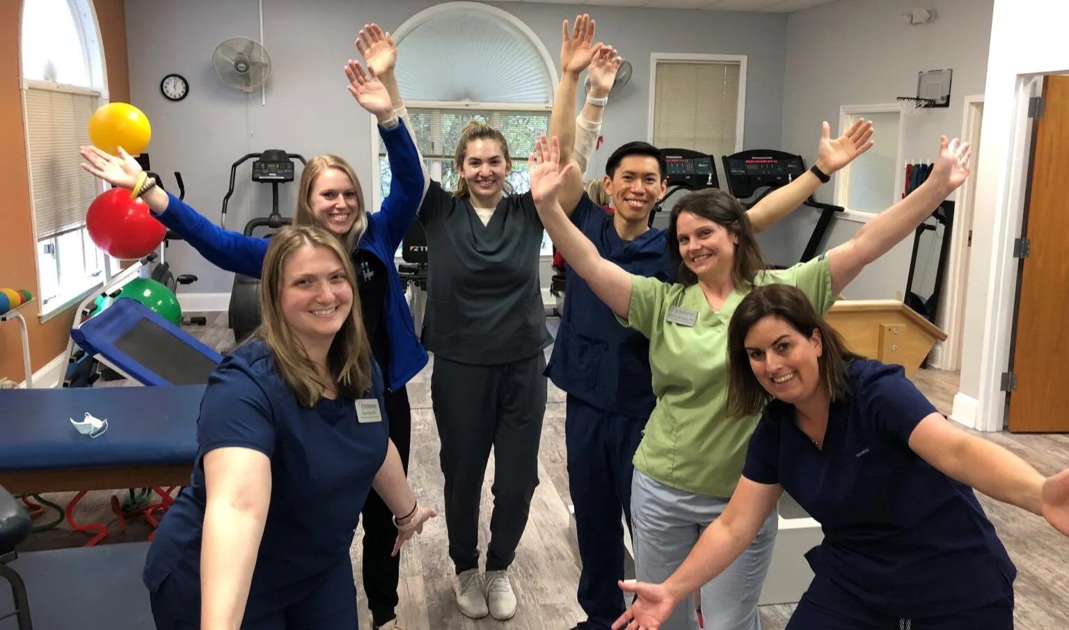 A group of six healthcare professionals in scrubs celebrates joyfully with arms raised, surrounded by exercise equipment in a therapy gym.